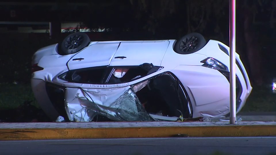 Imagen de WSVN 7News del sedán blanco volcado tras el choque de motocicleta en Hillsboro Boulevard en Deerfield Beach