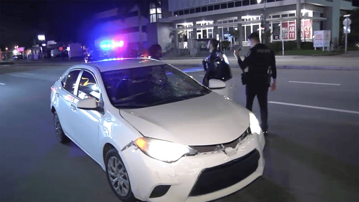 Escena nocturna de NBC 6 de un sedán blanco con daños en el parabrisas y el frente en North Federal Highway en Hallandale Beach, con luces de policía y agentes (sin rostros de víctimas).